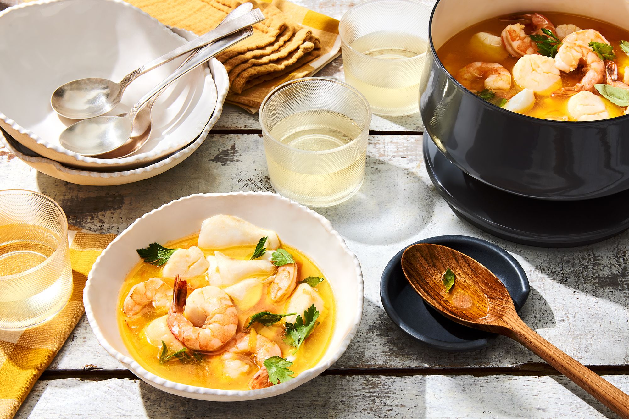 A small pot and bowl of seafood stew next to more bowls, serving utensils, wine, and napkins.